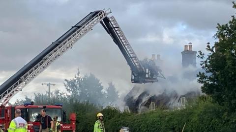A thatched building smoking with firefighters using a crane to get in close to the structure. The sky is filled with grey smoke. The roof of the building appears to have burned completely to nothing.