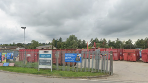 Entrance of Purton HRC. Lots of large red sorting containers around a concrete concourse. The sign at the entrance says HILLS, Wiltshire Council.