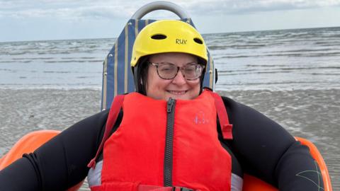 Naomi Herron in a floating chair with orange arms and blue back. She is wearing a black wetsuit, a red lifejacket, and a yellow hat. She is wearing glasses. She is on the beach.