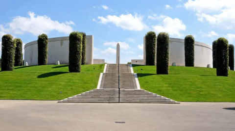 Neat and tidy grass and cropped trees surround a long, sweeping concrete pathway up to a memorial at the site. There is a circular design to it with a large monument or plinth in the middle.