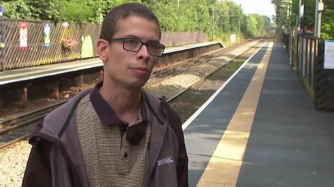 Nathaniel Yates is standing on the platform next to the railway line at Reddish North station. He is wearing glasses with a patterned shirt and a grey-and-black jacket. 