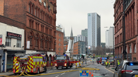 An image taken from street level of two fire engines with yellow hoses laid across the road. A grey undercover police vehicle is positioned to the right of the image next to a blue sign that blocks the road. Old red brick gothic buildings are on either side of the street. 