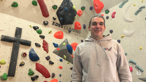 A man with dark hair and a light grey top is standing in front of a white wall with multi-coloured shapes on it.