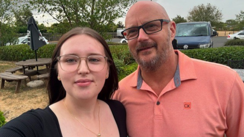 A selfie of a young woman with long dark hair, a black top and light brown glasses, and a bald man with black glasses and grey facial hair wearing a salmon pink t-shirt