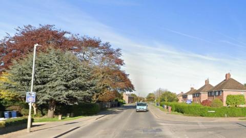 A quiet-looking road, bordered by 1950s houses and large trees. There is a junction on the right.