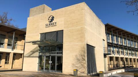 A sand-coloured building with a glass door and awning. The building has a sign reading "Surrey County Council" and a logo of a leaf above the awning.