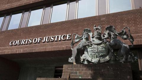 A brown-bricked building with a statue of a coat of arms on a plinth outside.