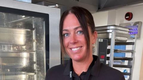 Woman with brown hair smiling at the camera with kitchen shelves behind her