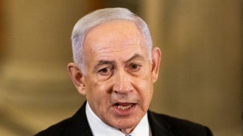 Israel's Prime Minister Benjamin Netanyahu wears a white shirt and black blazer, gazing slightly below the camera mid speech. 