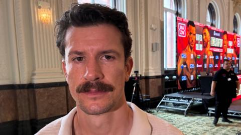 Boxer Nathan Heaney, who has dark hair and a moustache, stands to the side of a promotional dais advertising his next bout. A security man stands in the background to his left