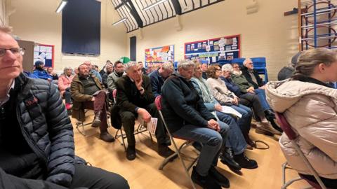 Rows of people are sat down on chairs in a public meeting. The setting is in a primary school hall, with a climbing frame on the wall and posters.