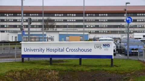 A sign in the foreground reads University Hospital Crosshouse. The grey hospital building is in the background with a red roof with several cars parked in front.