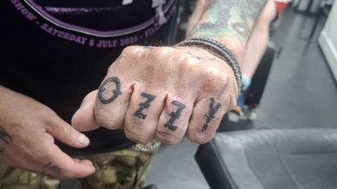 A man posing with his arm toward the camera. He is wearing a black and purple Black Sabbath concert t shirt. His hand is close to the camera, with a closed fist showing his Ozzy tattoo across his four fingers