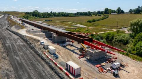 Red section of viaduct decking along the HS2 route, which shows as a light brown track on the ground. The decking is supported by concrete pillars. There is a darker area soil to the left of the track, and green fields with trees to the right.