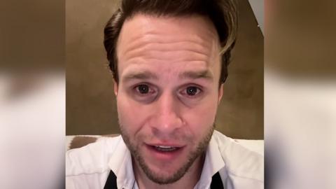 Olly Murs looks directly at the camera, he wears a white shirt and black tie