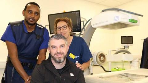 Keiran is seated in front of two clinicians. Keiran is wearing a black hoodie over a black t-shirt. The male clinician standing behind Keiran is wearing a blue protective scanning apron, while a female member of staff is standing with her hand on Keiran's left shoulder. All three are in front of a scanning machine.