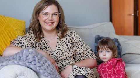 A woman with a spotty top sits on a sofa next to a child in a red outfit