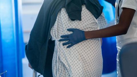 A health worker wearing a blue rubber glove with their hand guiding the back of a patient wearing a white and blue hospital gown.