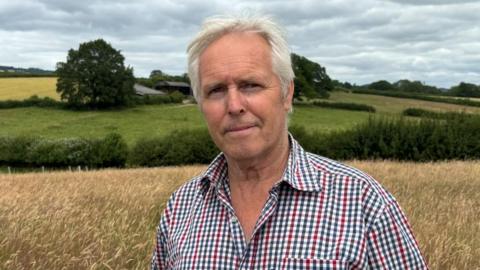 There is a man with light white hair, brushed back. He has a light stubbly beard. He has a blue, red and grey chequered t-shirt, with white squares in the middle. He has two buttons undone. Behind him is yellow grazing land, a dark green hedgerow and fields behind him.
