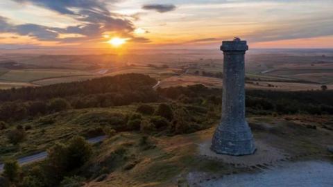 A tall grey octagonal brick tower stands to the right of the picture overlooking rolling countryside. A sunset on the horizon has turned the sky orange and pink.