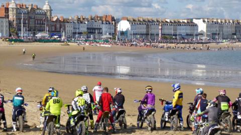 Line of motocross riders on sandby beach waiting to race along the sands with the sea to the right and seafront buildings to the left.
