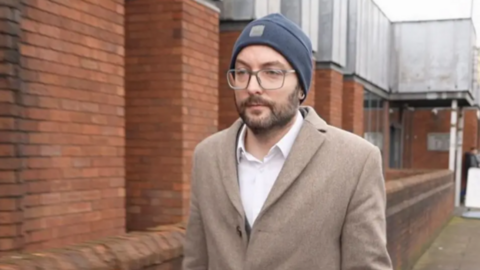 pencer is walking down a street and is wearing a brown coat a white shirt and a grey hat. He is also wearing glasses and has a brown beard.