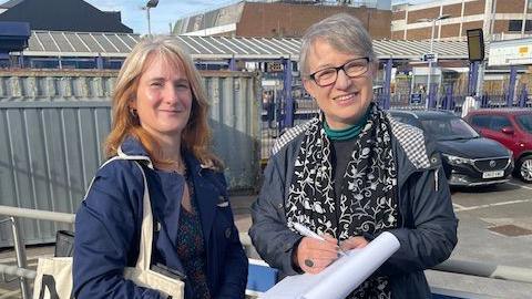 A woman in a blue coat and a white tote bag is standing next to another woman in a dark grey coat. She is holding a pen and signing a petition.