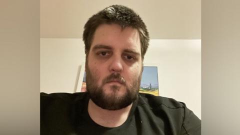 A man with dark hair and a dark beard looks into the camera. He is wearing a black t-shirt. 