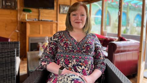 Lorna smiles at the camera as she sits on a chair with a sofa and a table behind her