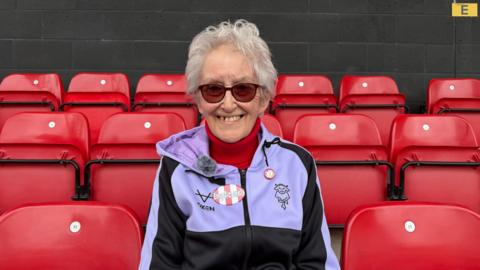 A woman sitting in one of the red seats at a football stadium. She has short grey hair and is smiling at the camera. She is wearing a black and purple zip-up hoodie with an Imps logo on it and two red and white pin badges. Underneath, she is wearing a bright red roll-neck jumper. She is also wearing sunglasses.