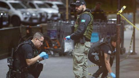 A Brazilian police officer kneeling down on the left of the picture takes samples at São Paulo airport. Another officer stands holding a see-through bag, while another officer on the right bends down to pick something up. 