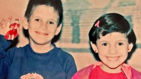 A boy in a blue top and a girl in a pink top, both with dark hair, smile at the camera - the boy is holding up a toy figurine