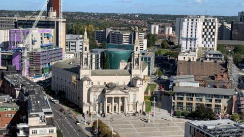Image shows an ariel shot of Leeds Civic Hall with Millenium Square. 
Behind is a Leeds University glass building with a sky line scene.
To the left of the shot is the Leeds General Infirmary.
