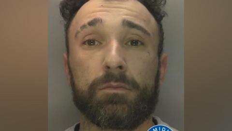 Cain Watson has a short brown beard and short, curly brown hair, as he stares at the camera in his mugshot.