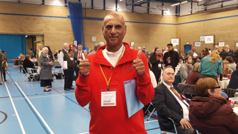 Milkinder is stood in a sports centre at a local election count giving the thumbs up with both thumbs. He has a red jumper on and brown trousers and a lanyard around his neck. There are many people standing and sitting in the centre behind him.