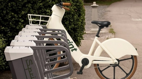 The picture shows a row of bike‑hire docking stands positioned along the edge of a path. One electric bike is locked into one of the stands. The bike is cream‑coloured with brown handle grips and tyres, a front basket and a solid rear wheel cover. The word “Sprint” is printed in green lettering along the bike’s frame and also appears on the side panels of the docking stands.