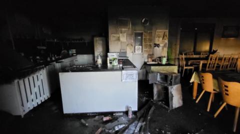 Inside burnt cafe, with extensive scorch marks and fire damage. A great deal of the contents is covered in soot. 