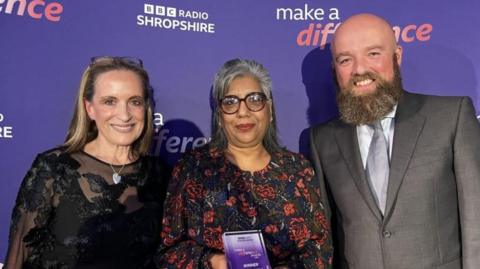 Two women and one man with one woman holding a small purple award. The two women have patterned black tops and the man is wearing a grey suit and has a beard