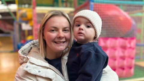 Carla Jones is holding her child Oakley and smiling at the camera. She is in front of a soft play area.