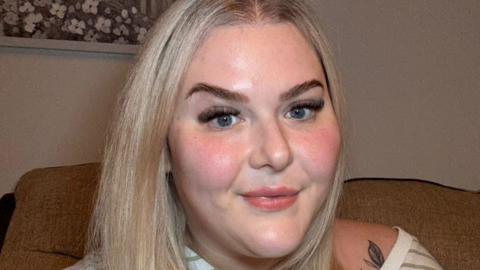 A woman with blonde hair smiles with closed lips as she looks at the camera. She is sitting on a beige sofa in a room and has a striped green and white top slipping off her shoulders