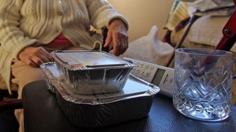 Two foil food containers stacked, and empty glass and a landline wireless phone sit on a black leather-like surface. Behind it is an elderly person wearing a beige cable knit cardigan with buttons. Face is not visible and clothing does not make it possible to distinguish if they are male or female.