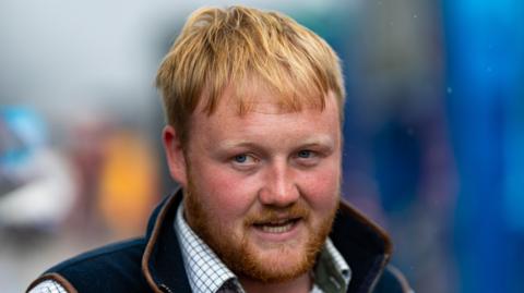Kaleb Cooper, a blonde man with a beard wearing a white and blue shirt under a gilet, looks into the distance with a blurred background behind him.