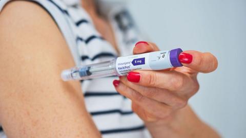 A woman with red nail polish is injecting herself in the arm. The needle is in a tube which is labelled "Mounjaro 5mg, Kwikpen". She is wearing a striped top.