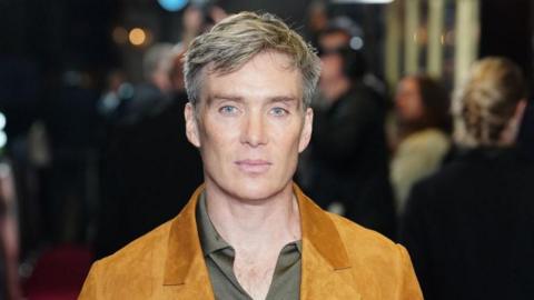 Cillian Murphy has short greyish hair and is wearing a mustard blazer and a light green shirt.