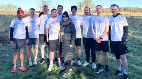 Steve has his arm around Ruth's shoulder. Steve and the team wear black shorts and white tops. Ruth wears a black puffer jacket with a hat and scarf.
