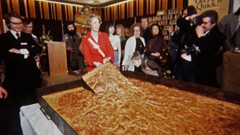 Woman in red dress lifting up a large slice of Yorkshire pudding from a huge baking tray, in front of press photographers.