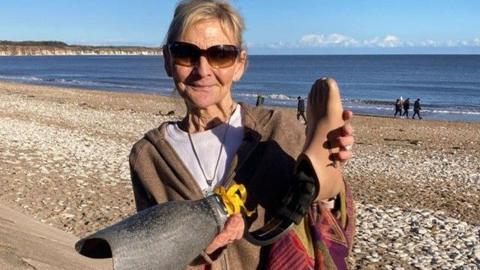 Brenda holds the prosthetic up - she is on the beach smiling.  The sun is out and she is wearing sun glasses.