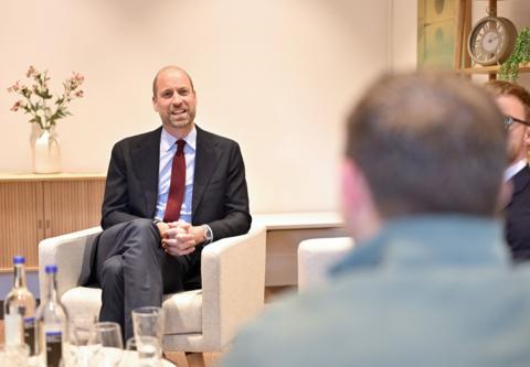 A balding man with a beard sat in a white armchair chatting to other men. He is wearing a blue blazer, grey trousers, blue shirt and claret tie