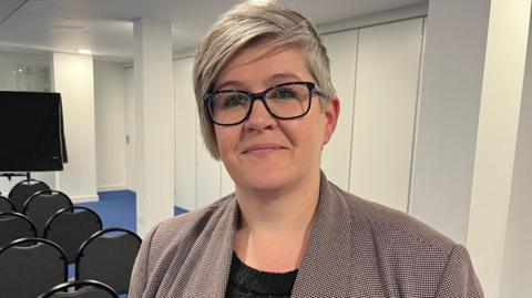 Deputy Haley Camp is pictured. She is a woman with short grey hair, wearing black glasses and a checked jacket on top of a black blouse. 