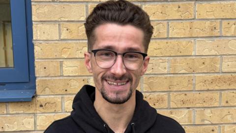 A headshot of a man standing in front of a bright wall. He has thick brown hair which is styled upwards and short on the sites. He has a goatee and wears thin-rimmed glasses. He is wearing a black hoodie.